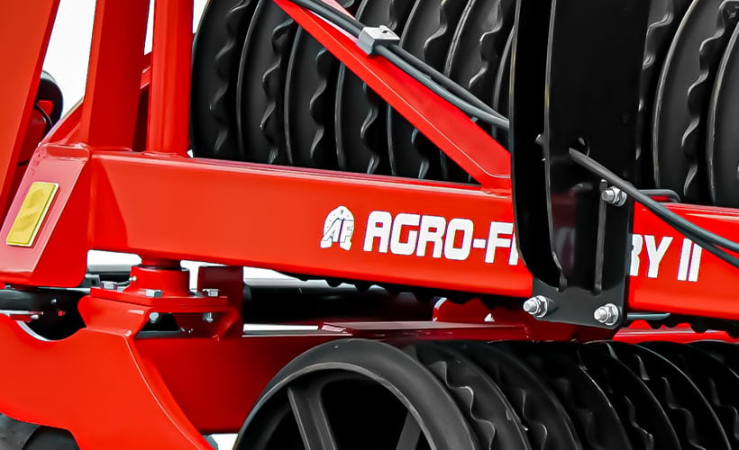 Part of a red agricultural roller from AGRO-FACTORY, with the company&apos;s logo visible on the side shield. The machine is equipped with dark, ringed rollers, which are connected to the main arm via black, metal connectors. Construction details such as screws, clamps, and fastening elements are clearly visible, emphasizing that the agricultural rollers of this brand are solid and durable farming machines.