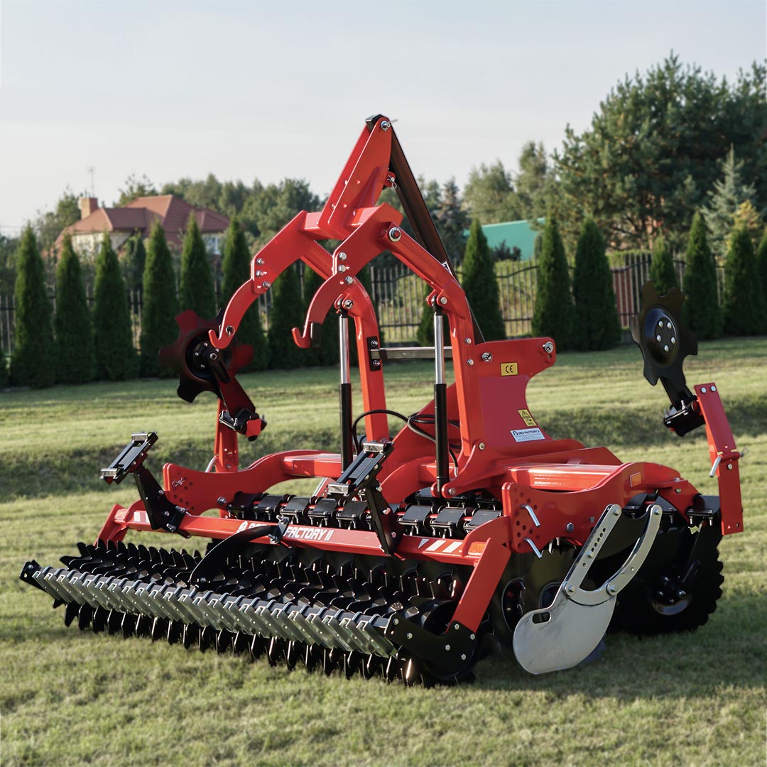 In the photo, a red THOR disc harrow from Agro-Factory is shown. These agricultural machines, used in farming, are designed for intensive soil cultivation, especially for loosening and mixing it with plant residues. Characteristic of this type of equipment are the sharp discs arranged in a row, which cut and mix the soil, preparing it for the next stages of cultivation. The solid construction with visible mounts and connectors, coated in red paint, indicates the durability of the device. The machine is also equipped with support wheels, which allow for easy maneuvering and transport in the field and between fields. The design of the aggregate is modern and functional, suggesting that the manufacturer focuses on providing farmers with high-performance and reliable machines. The placement of the machine against a backdrop of green grass and trees, near residential buildings, may suggest that it is suitable equipment for both larger and smaller farms, whose owners value both the aesthetics and functionality of their equipment.