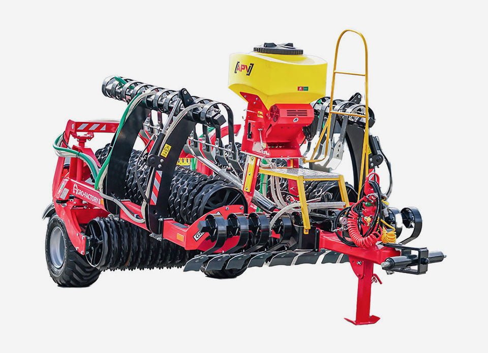 A modern, red agricultural roller from Agro-Factory with yellow and black additions, set against a white background. The machine is equipped with a yellow APV seeder mounted on top, a comprehensive hydraulic system with red and black hoses, and ring rollers, as well as yellow steps and handrail ensuring safe access. The entire unit presents itself as highly advanced farming equipment for precise soil cultivation.